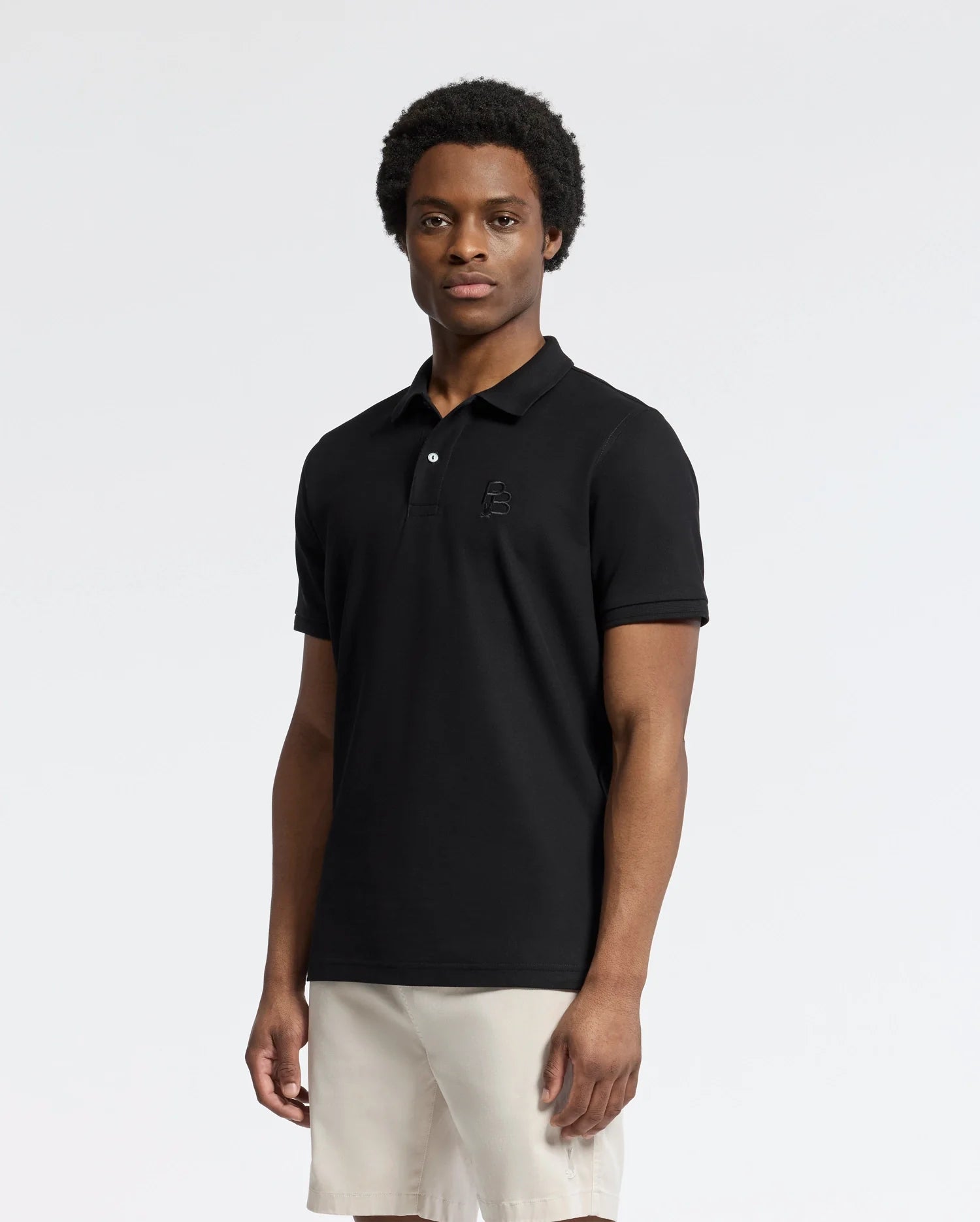 A man wears the Psycho Bunny Men's Nicholson Rib Collar Pique Polo (B6K401F200) in classic black with an embroidered logo, paired with beige shorts, crafted from soft Pima cotton, against a plain, light background.