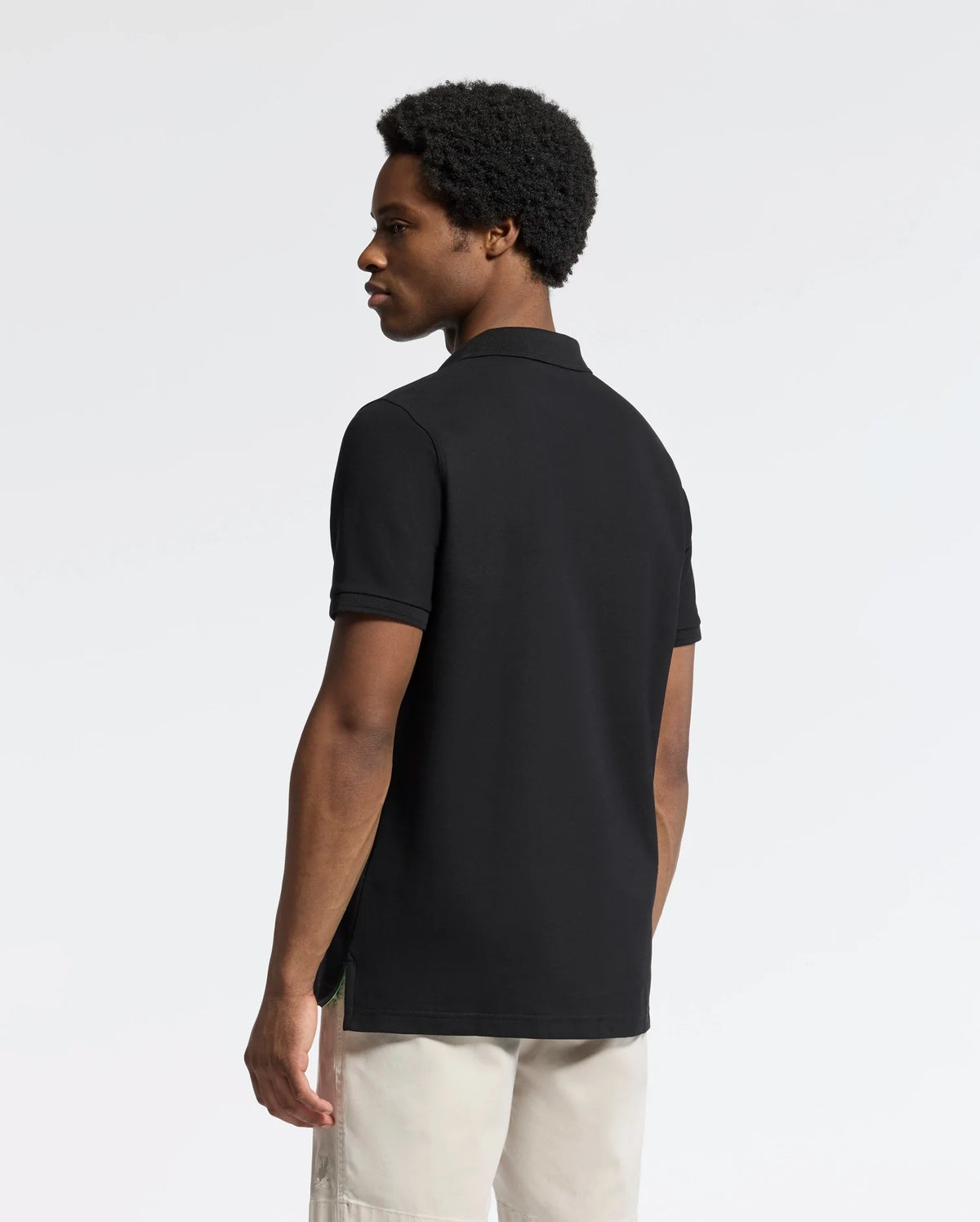 A person with short curly hair stands slightly turned away, wearing light-colored pants and a classic black MENS NICHOLSON RIB COLLAR PIQUE POLO by Psycho Bunny made of Pima cotton. The plain, light background subtly enhances the embroidered logo on their polo.