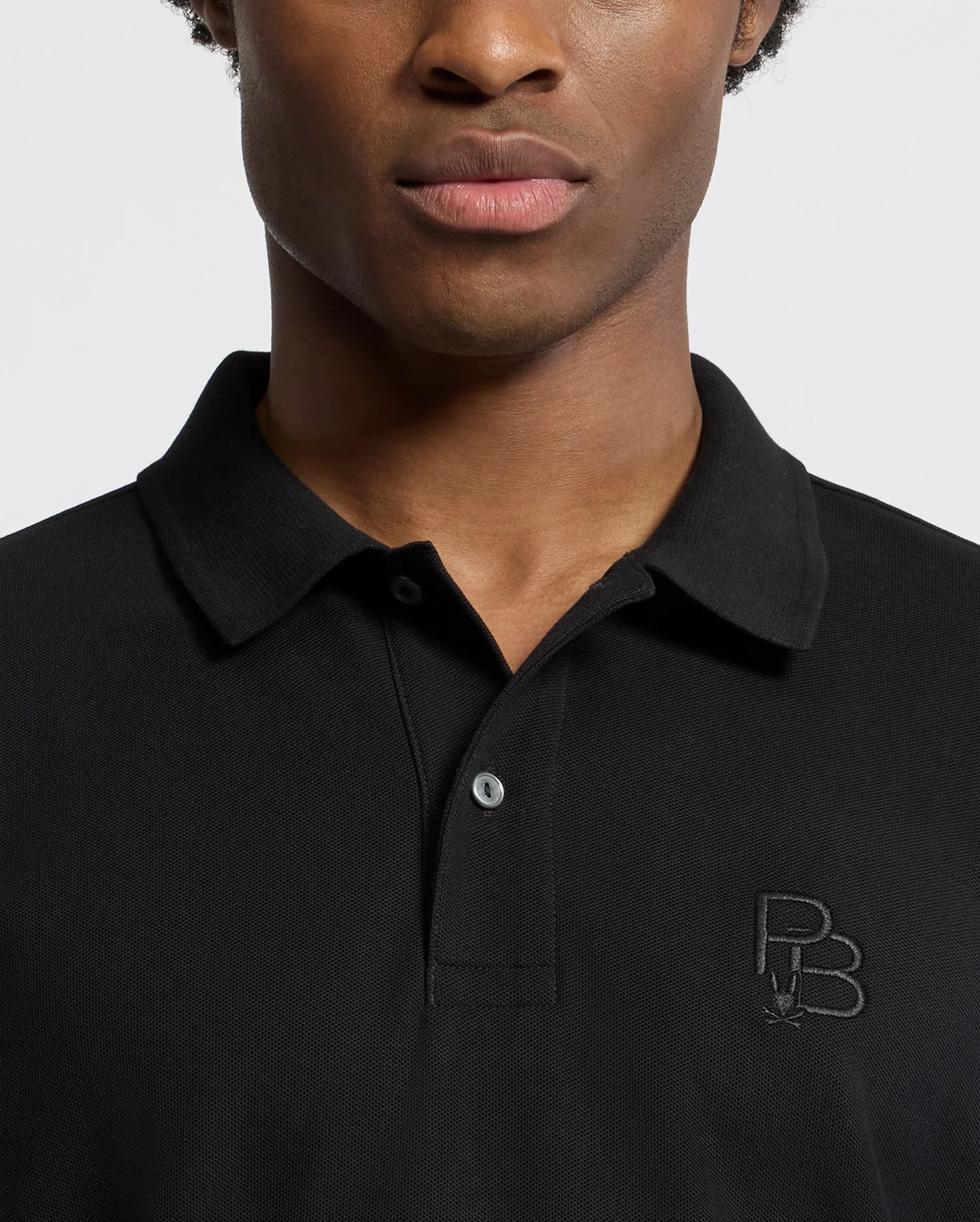 A person wearing the Psycho Bunny MENS NICHOLSON RIB COLLAR PIQUE POLO - B6K401F200 in classic black, made from soft Pima cotton, with "PB" initials embroidered on the left side. The short sleeves and undone top button create a relaxed yet refined look against the plain white background.