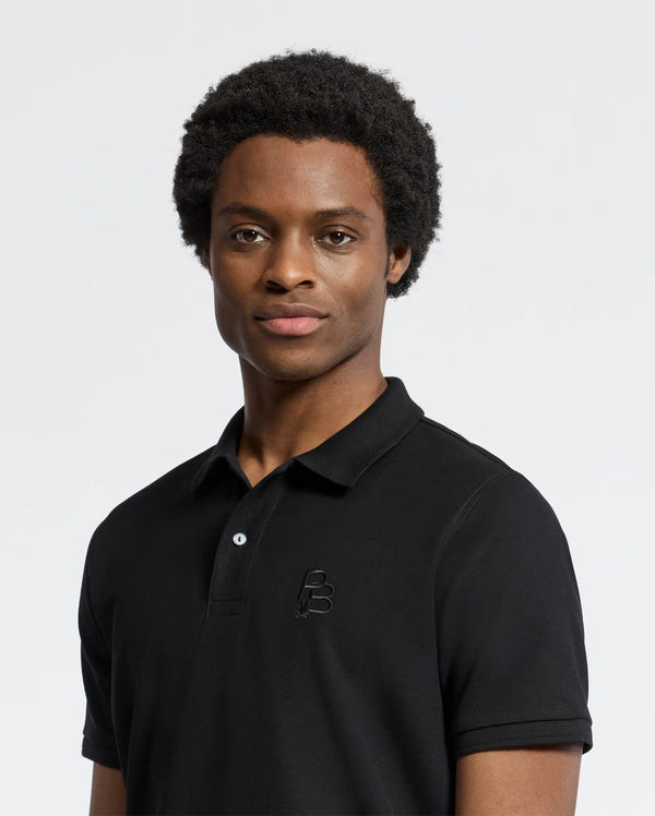 A man with short curly hair wears the Psycho Bunny Men's Nicholson Rib Collar Pique Polo (B6K401F200) in classic black, made from soft Pima cotton with a subtle embroidered logo. He stands against a light background, looking at the camera neutrally.