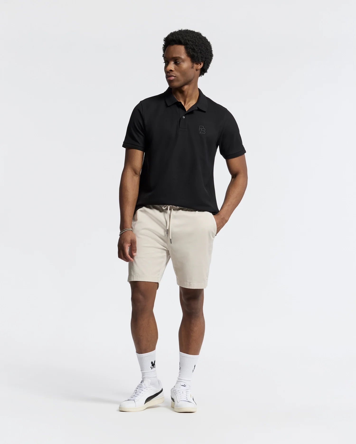 Against a white background, a person wears the MENS NICHOLSON RIB COLLAR PIQUE POLO by Psycho Bunny in black, paired with beige shorts, white socks, and sneakers. They have short curly hair and gaze to their right.