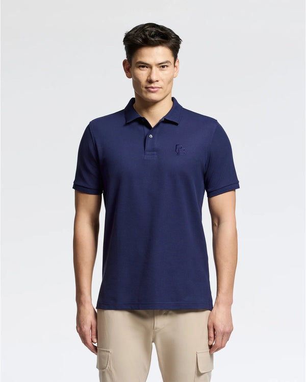A man stands against a plain white background, wearing the Psycho Bunny MENS NICHOLSON RIB COLLAR PIQUE POLO - B6K401F200 in navy blue, made of soft Pima cotton with Mother-of-Pearl buttons and a small embroidered logo on the left chest. Paired with beige pants, he looks directly at the camera.