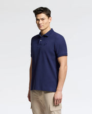 A man wears the Psycho Bunny MENS NICHOLSON RIB COLLAR PIQUE POLO in dark blue, featuring an embroidered chest logo and made from Pima cotton. He pairs it with light-colored cargo pants against a plain background.