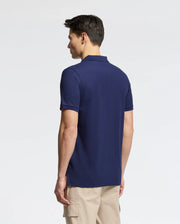 A person with short dark hair faces away from the camera, wearing a Psycho Bunny Men's Nicholson Rib Collar Pique Polo in dark navy blue with Mother-of-Pearl buttons and light beige pants. The background is plain and neutral.