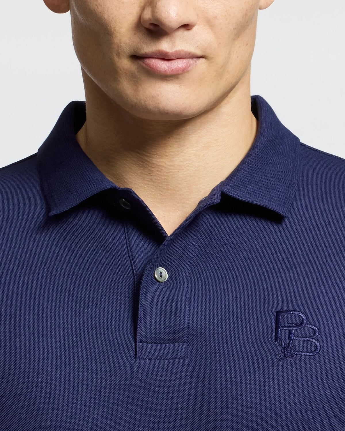 A person is wearing a Psycho Bunny MENS NICHOLSON RIB COLLAR PIQUE POLO (B6K401F200) in navy blue, made from soft Pima cotton with a subtle chest logo. The classic collar and elegant two Mother-of-Pearl buttons stand out against the light gray background.