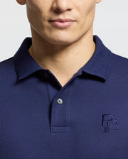 A person is wearing a Psycho Bunny MENS NICHOLSON RIB COLLAR PIQUE POLO (B6K401F200) in navy blue, made from soft Pima cotton with a subtle chest logo. The classic collar and elegant two Mother-of-Pearl buttons stand out against the light gray background.