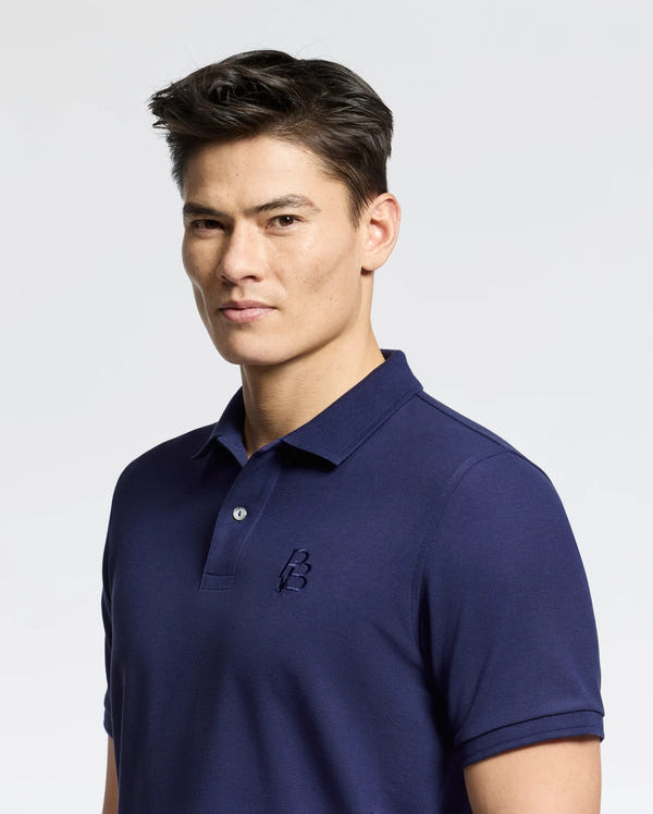 A person with short dark hair wearing a navy Psycho Bunny Nicholson rib collar pique polo, featuring mother-of-pearl buttons and a small embroidered "B" on the chest, stands against a plain light background.