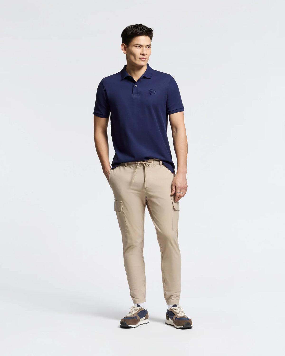 A man in a Psycho Bunny MENS NICHOLSON RIB COLLAR PIQUE POLO stands against a plain white backdrop. He's wearing beige cargo jogger pants and multicolored sneakers, with hands in pockets, looking to the side.