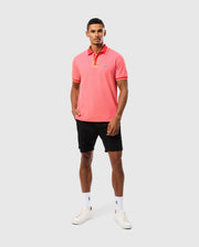 A person stands against a plain background wearing a MENS WOODWAY OXFORD POLO (B6K405C200) by Psycho Bunny, featuring a pink collared shirt with a classic two-button placket and an embroidered Bunny logo, black shorts, and white sneakers. The Pima cotton-blend fabric of the shirt complements their neutral expression as they have one hand in their pocket.