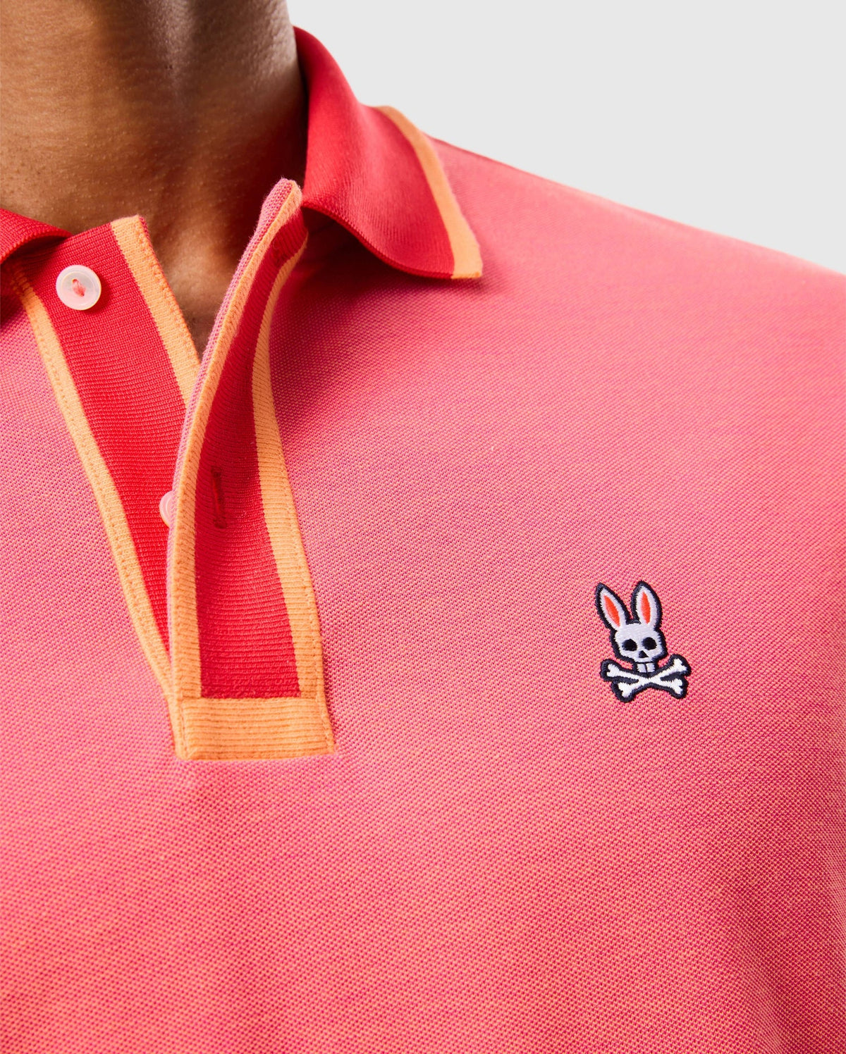 A close-up of a person wearing the MENS WOODWAY OXFORD POLO - B6K405C200 from Psycho Bunny, featuring a pink polo shirt with orange trim and white buttons. The classic two-button placket design is crafted from a Pima cotton-blend and showcases an embroidered Bunny logo with crossed bones on the chest. The background is plain and light-colored.