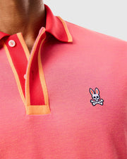 A close-up of a person wearing the MENS WOODWAY OXFORD POLO - B6K405C200 from Psycho Bunny, featuring a pink polo shirt with orange trim and white buttons. The classic two-button placket design is crafted from a Pima cotton-blend and showcases an embroidered Bunny logo with crossed bones on the chest. The background is plain and light-colored.