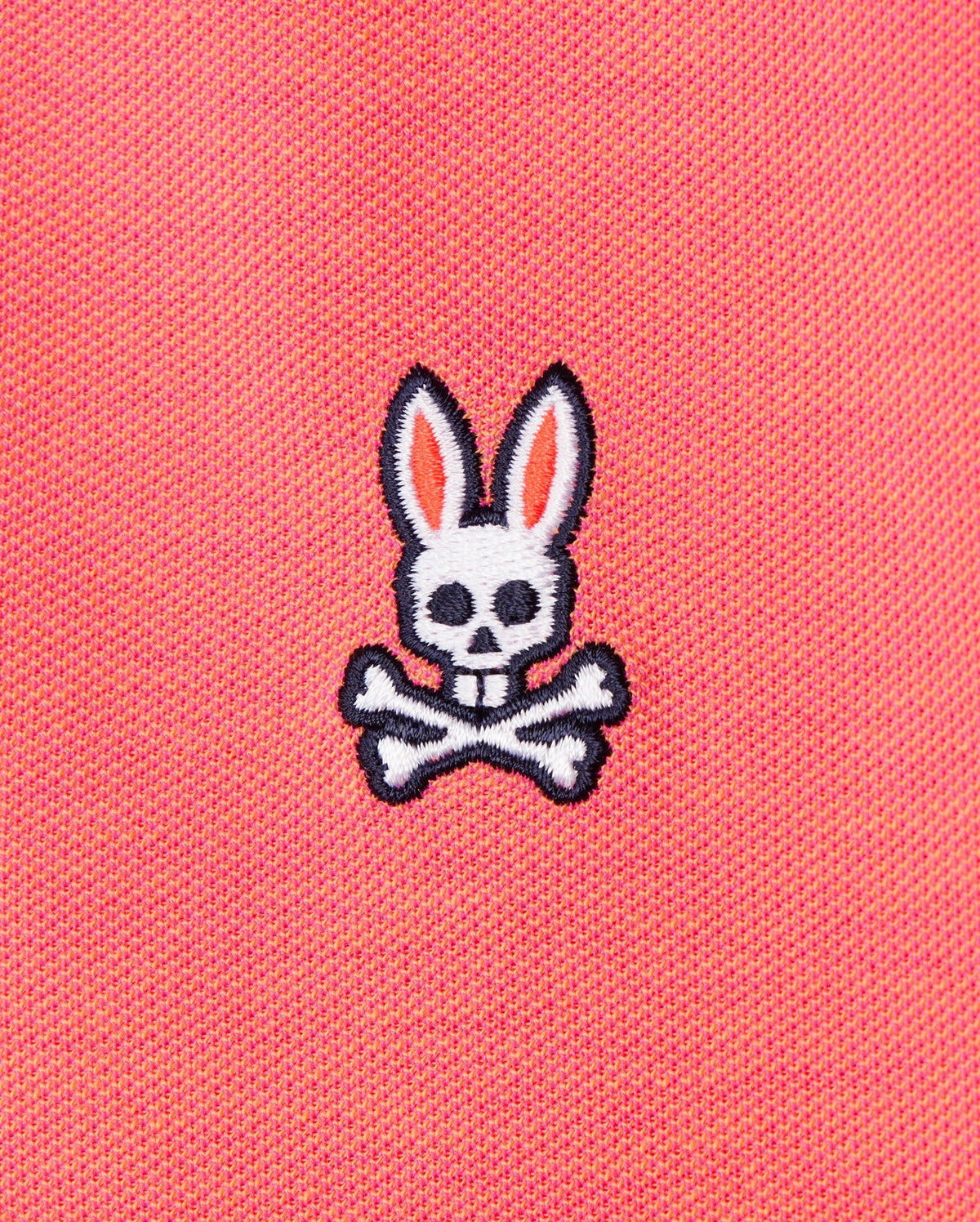 The image shows the Psycho Bunny MENS WOODWAY OXFORD POLO - B6K405C200, featuring a pink Pima cotton-blend fabric with an embroidered design of a white skull with rabbit ears and crossed bones underneath. The skull has black eye holes and a nose hole, while the ears are outlined in black with pink inner details.