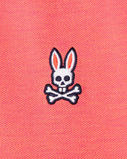 The image shows the Psycho Bunny MENS WOODWAY OXFORD POLO - B6K405C200, featuring a pink Pima cotton-blend fabric with an embroidered design of a white skull with rabbit ears and crossed bones underneath. The skull has black eye holes and a nose hole, while the ears are outlined in black with pink inner details.
