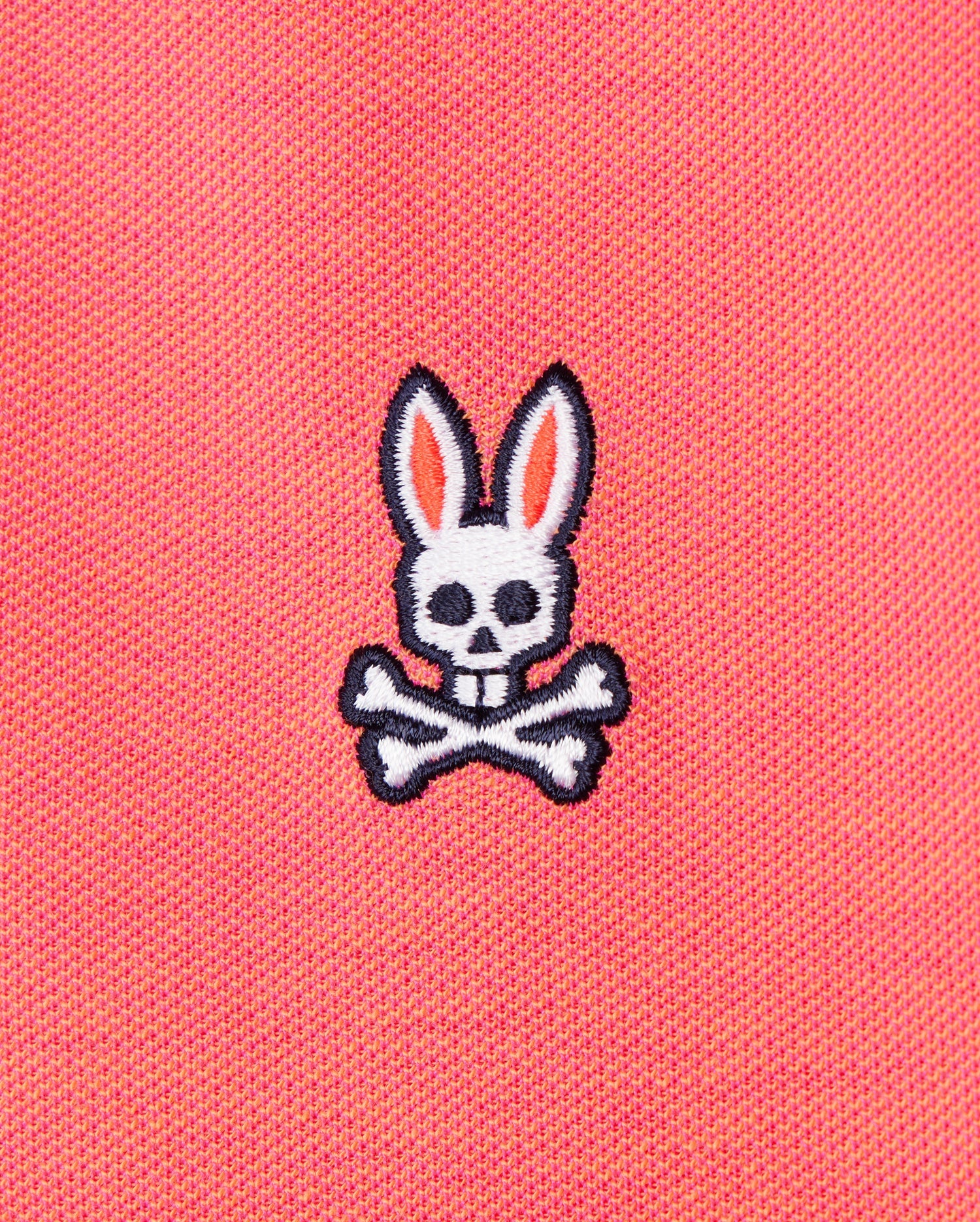The image shows the Psycho Bunny MENS WOODWAY OXFORD POLO - B6K405C200, featuring a pink Pima cotton-blend fabric with an embroidered design of a white skull with rabbit ears and crossed bones underneath. The skull has black eye holes and a nose hole, while the ears are outlined in black with pink inner details.