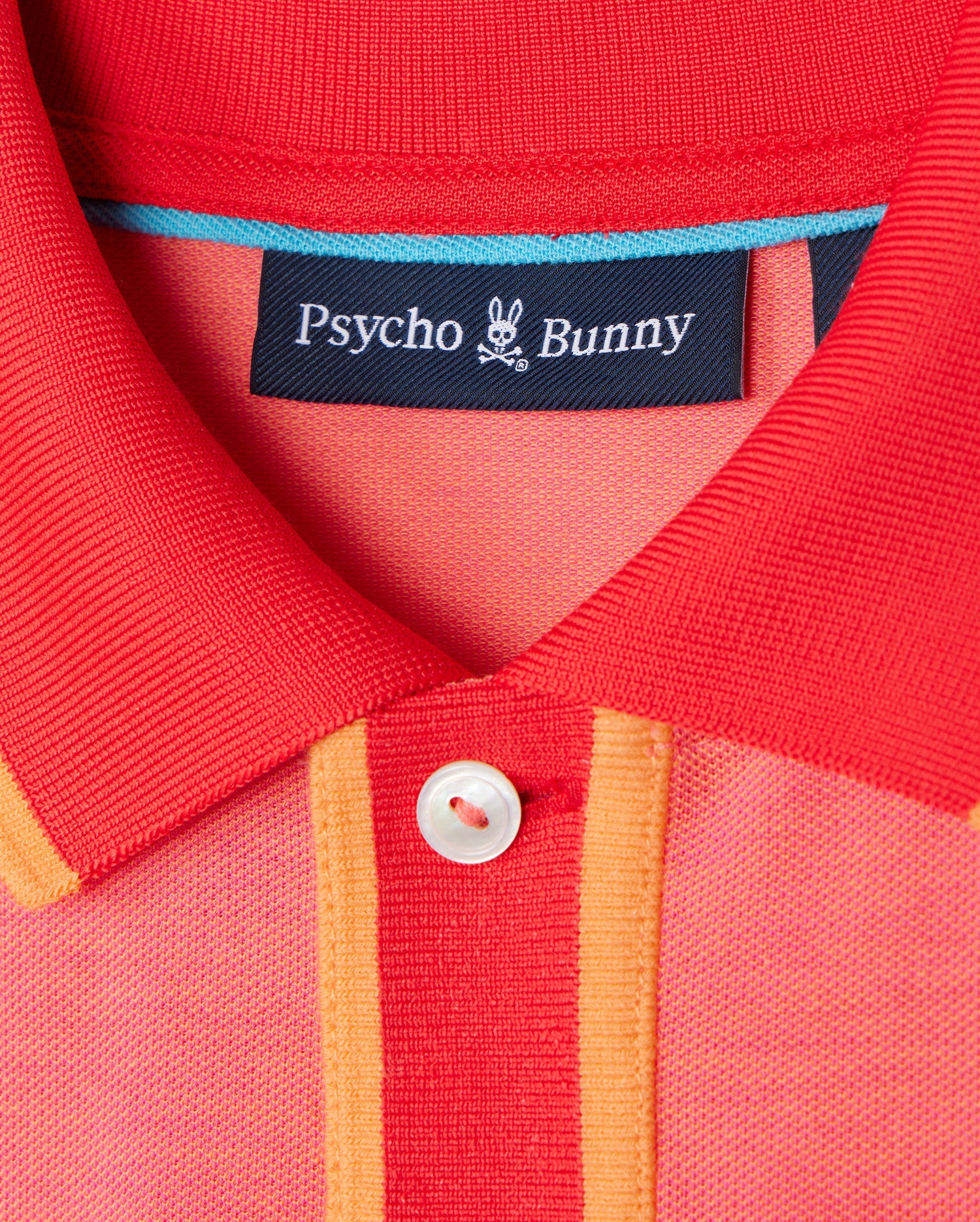 Close-up of the *Mens Woodway Oxford Polo - B6K405C200*, a red and orange collared shirt from Psycho Bunny. The label "Psycho Bunny" is attached to the inside of the collar. Made from a Pima cotton-blend, it features a classic two-button placket and a white button that contrasts with the vibrant fabric colors.