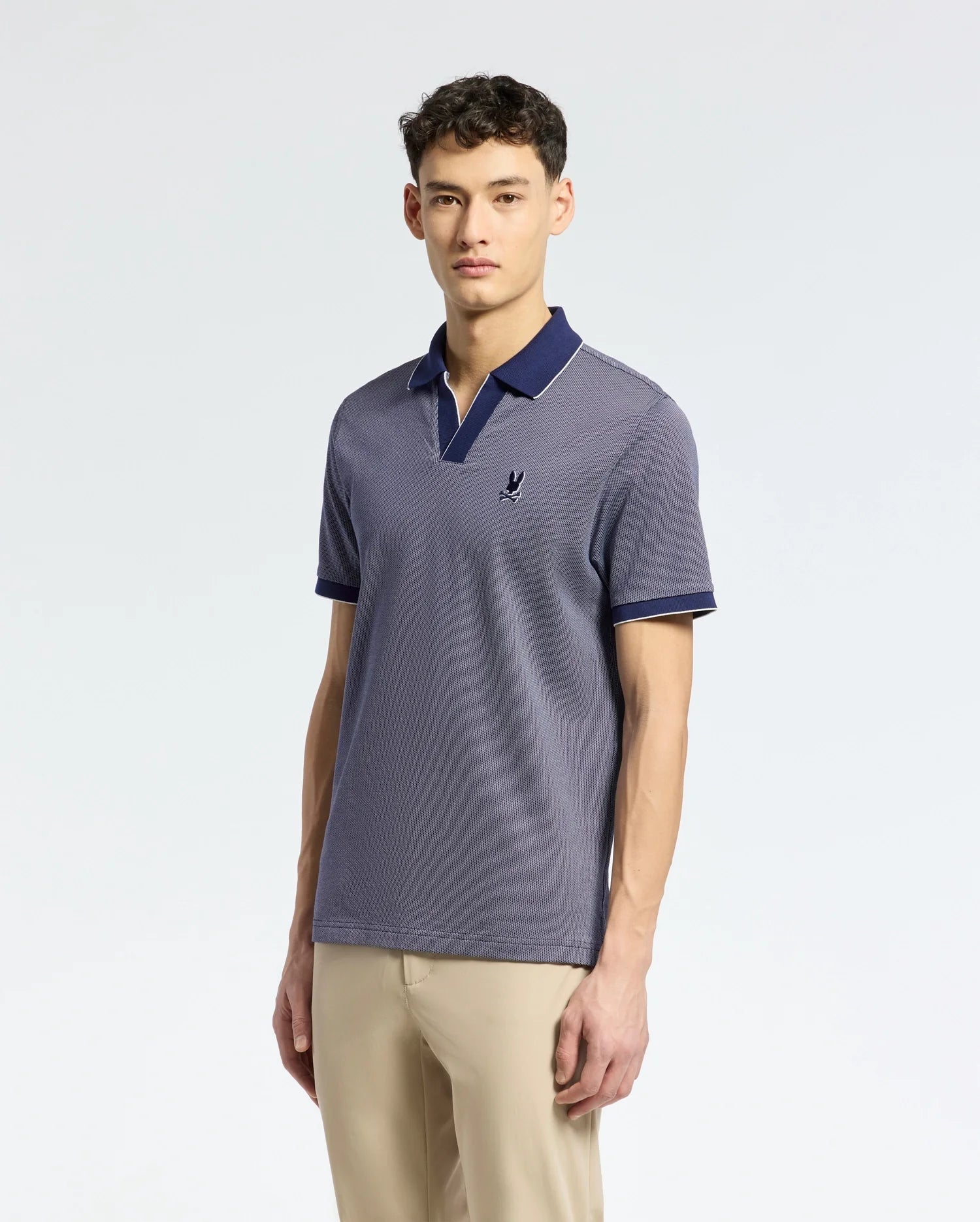 A person models Psycho Bunny's Men's Mackay Jacquard Johnny Collar Polo, B6K408F200, in light navy Pima cotton with a chest emblem and dark navy collar and sleeve edges, paired with beige pants against a plain backdrop.