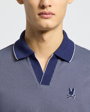 A person is wearing a Psycho Bunny blue Johnny-collared polo (MENS MACKAY JACQUARD JOHNNY COLLAR POLO - B6K408F200) in soft Pima cotton. It has white edging and a small embroidered rabbit with crossed bones on the chest, set against a plain white background.