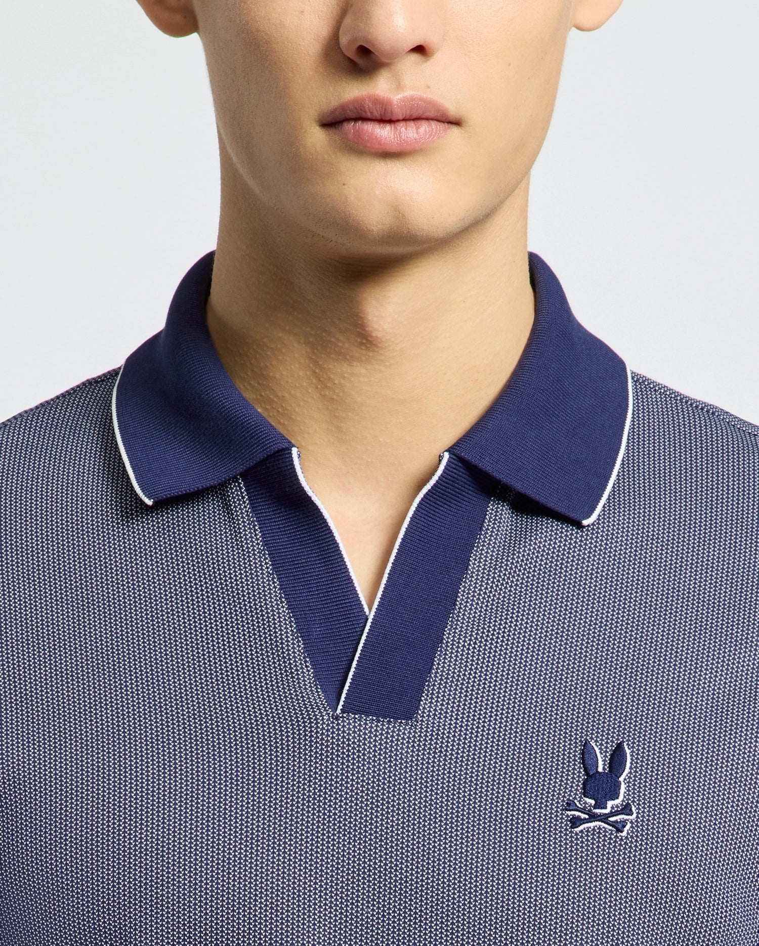 A person is wearing a Psycho Bunny blue Johnny-collared polo (MENS MACKAY JACQUARD JOHNNY COLLAR POLO - B6K408F200) in soft Pima cotton. It has white edging and a small embroidered rabbit with crossed bones on the chest, set against a plain white background.