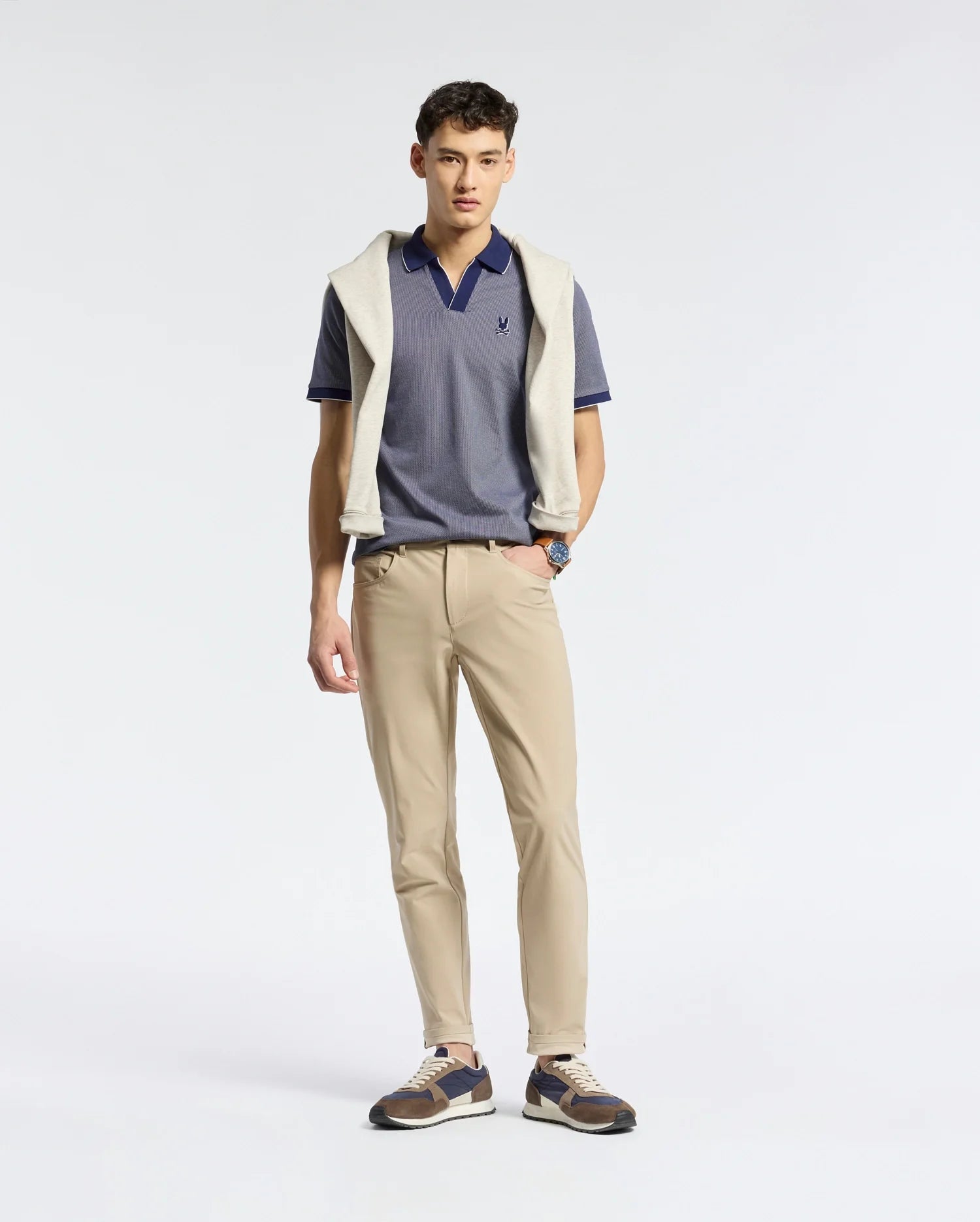 A man in a Psycho Bunny MENS MACKAY JACQUARD JOHNNY COLLAR POLO and beige pants stands against a plain backdrop, with a light sweater draped over his shoulders. He’s wearing sneakers, a wristwatch, and exudes a relaxed pose.