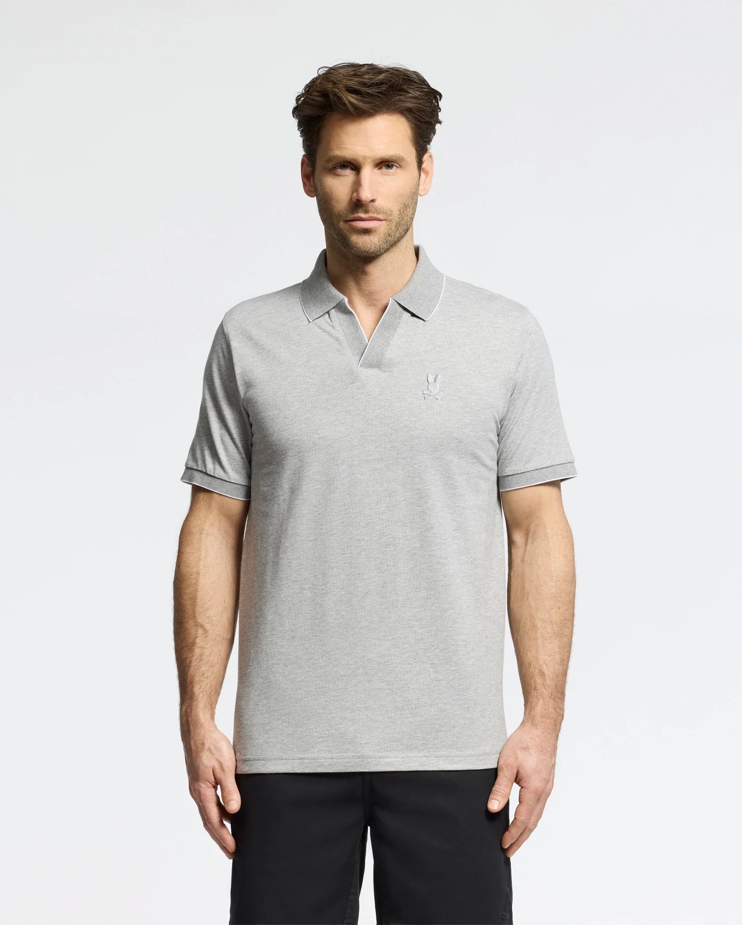 A man in a Psycho Bunny MENS MACKAY JACQUARD JOHNNY COLLAR POLO - B6K408F200, crafted from Pima cotton in light gray with a chest logo, stands against a white background. The polo pairs well with his dark pants. He has short brown hair and a neutral expression.