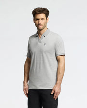A man with short hair wears the Psycho Bunny MENS MACKAY JACQUARD JOHNNY COLLAR POLO in light gray, featuring a small left chest emblem and classic collar, paired with black pants. He stands against a plain white background, looking directly at the camera with a neutral expression.