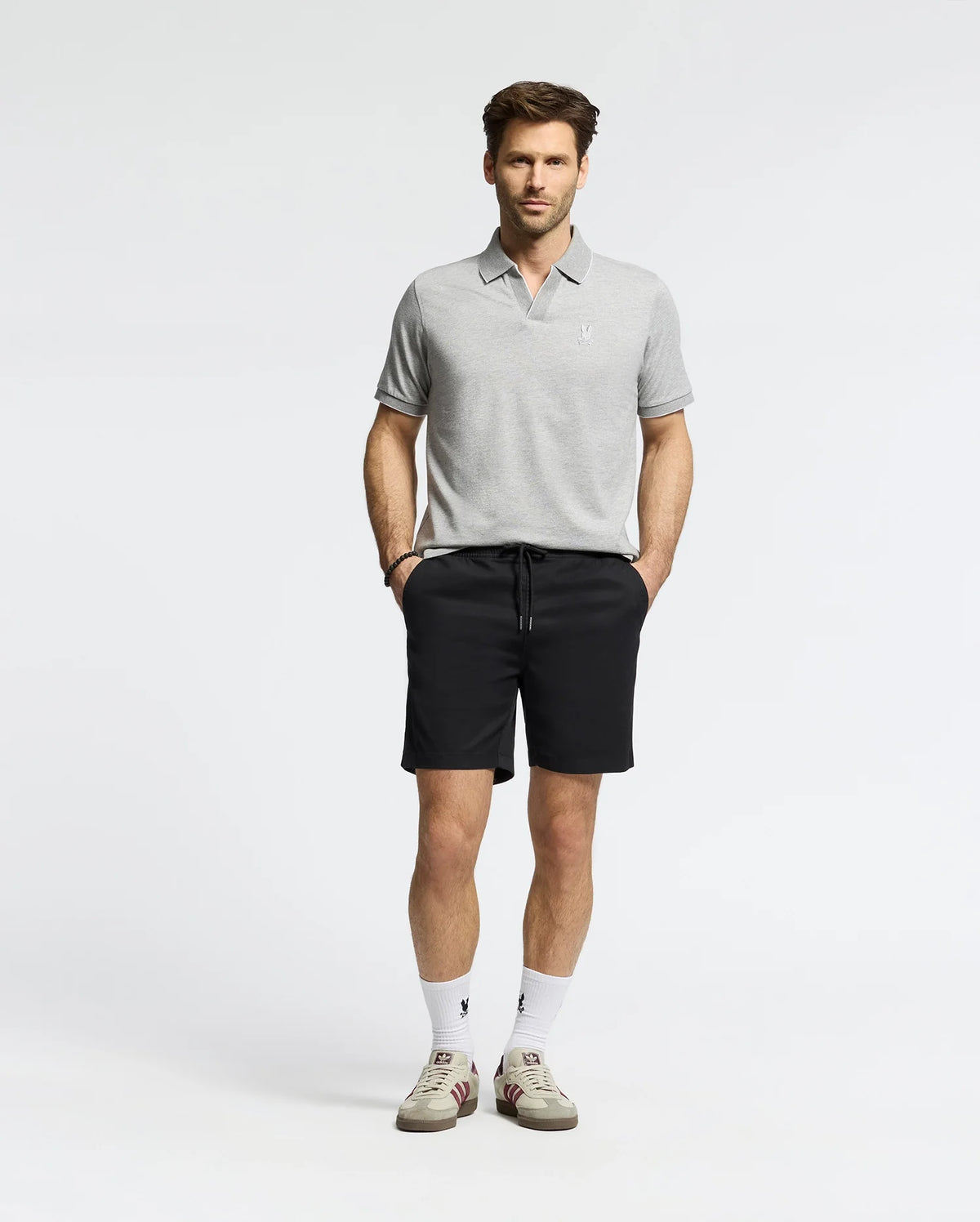 A man wears a Psycho Bunny MENS MACKAY JACQUARD JOHNNY COLLAR POLO (B6K408F200) in gray with black shorts, white socks, and pima cotton sneakers featuring a black "V" logo. He stands against a plain background with hands in pockets and a neutral expression.