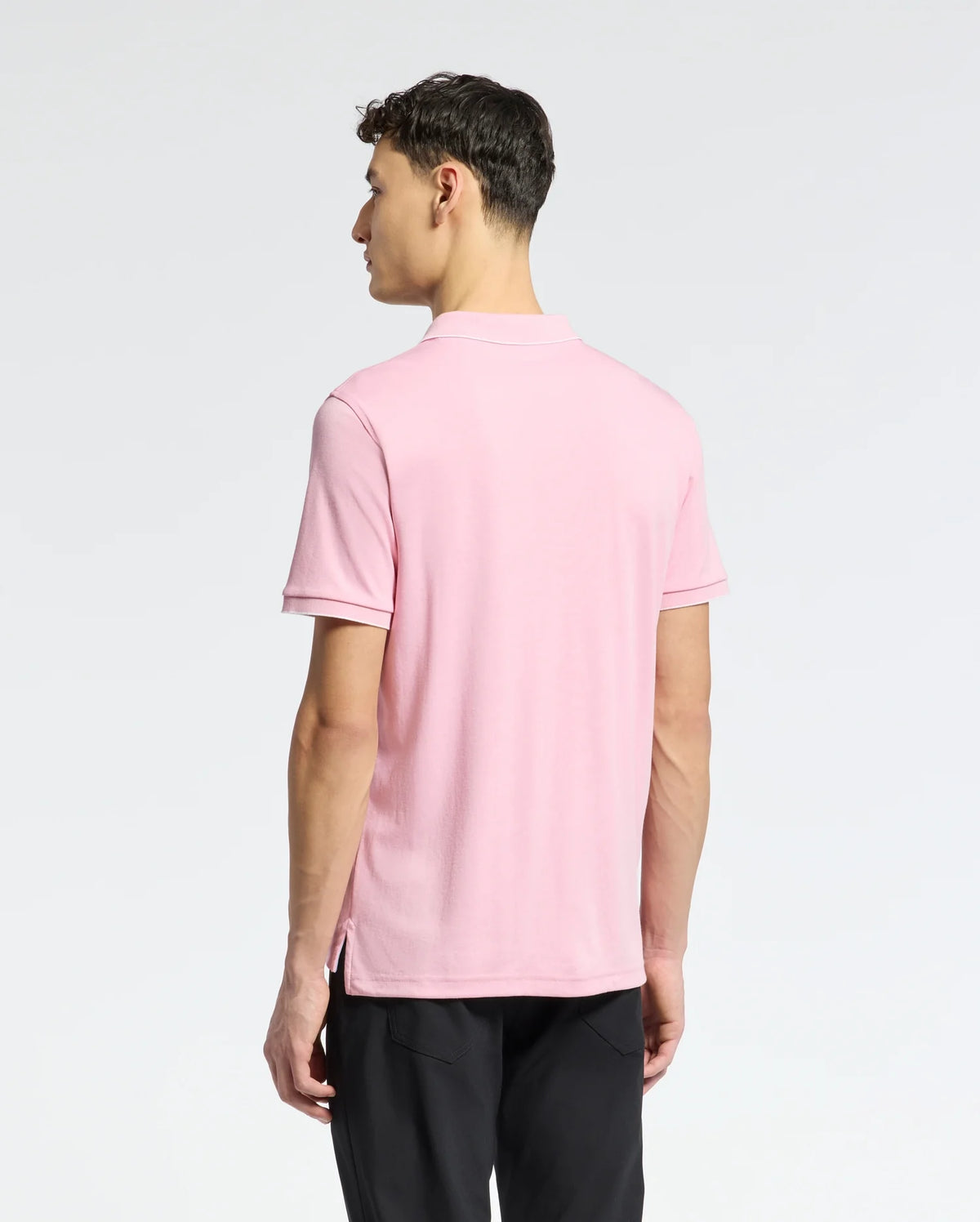 A person facing away from the camera is wearing a light pink Psycho Bunny MENS BRYCE PERFORMANCE PIQUE POLO (B6K486F200) and black pants. The plain light gray background highlights the sophistication of this moisture-wicking attire, perfect for active days.