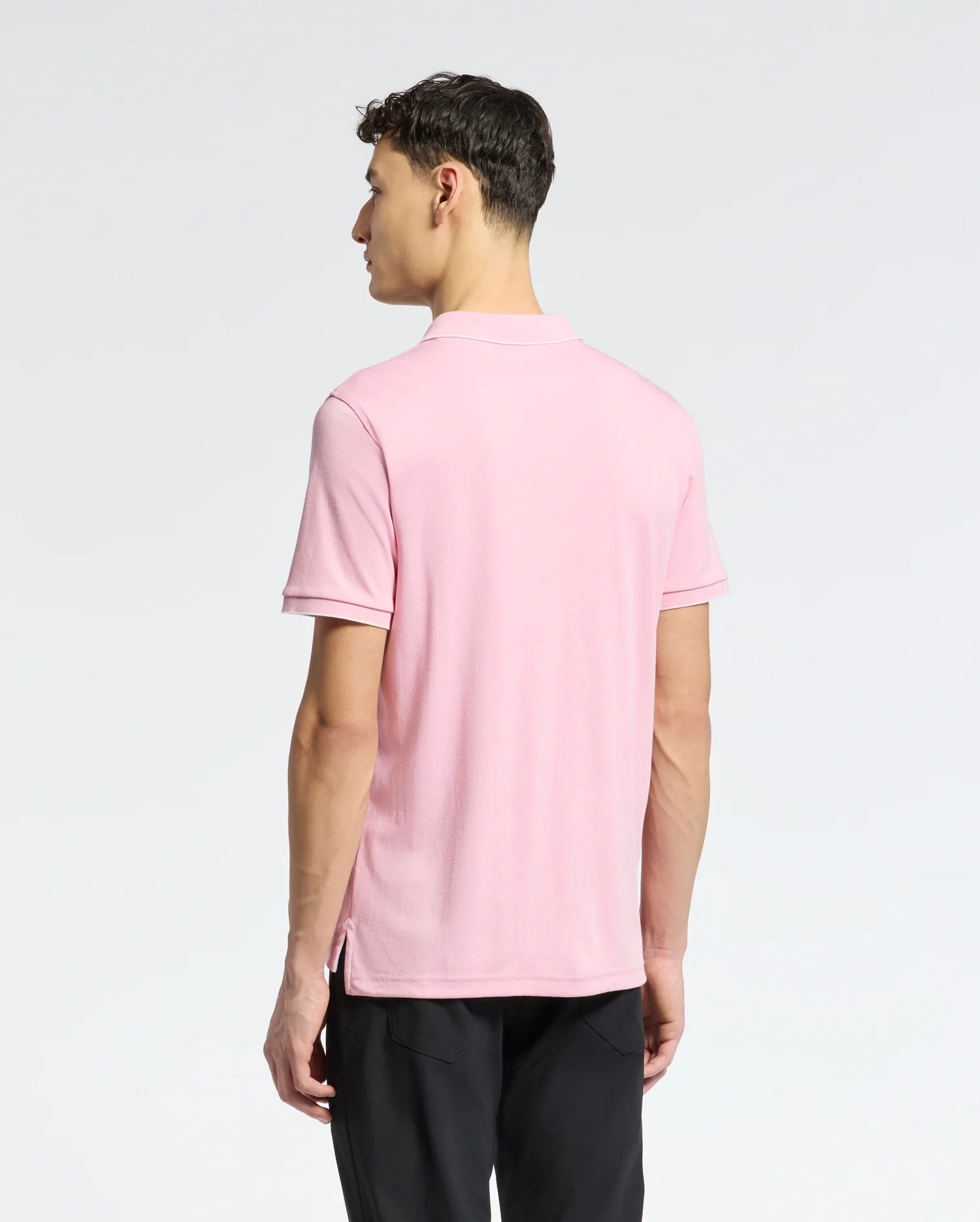 A person facing away from the camera is wearing a light pink Psycho Bunny MENS BRYCE PERFORMANCE PIQUE POLO (B6K486F200) and black pants. The plain light gray background highlights the sophistication of this moisture-wicking attire, perfect for active days.