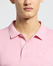 A person wearing a Psycho Bunny MENS BRYCE PERFORMANCE PIQUE POLO - B6K486F200 in light pink, featuring a small bunny logo on the chest. Made from performance pique fabric with quick-dry and moisture-wicking properties, it has a collar and two buttons. The background is plain and neutral.