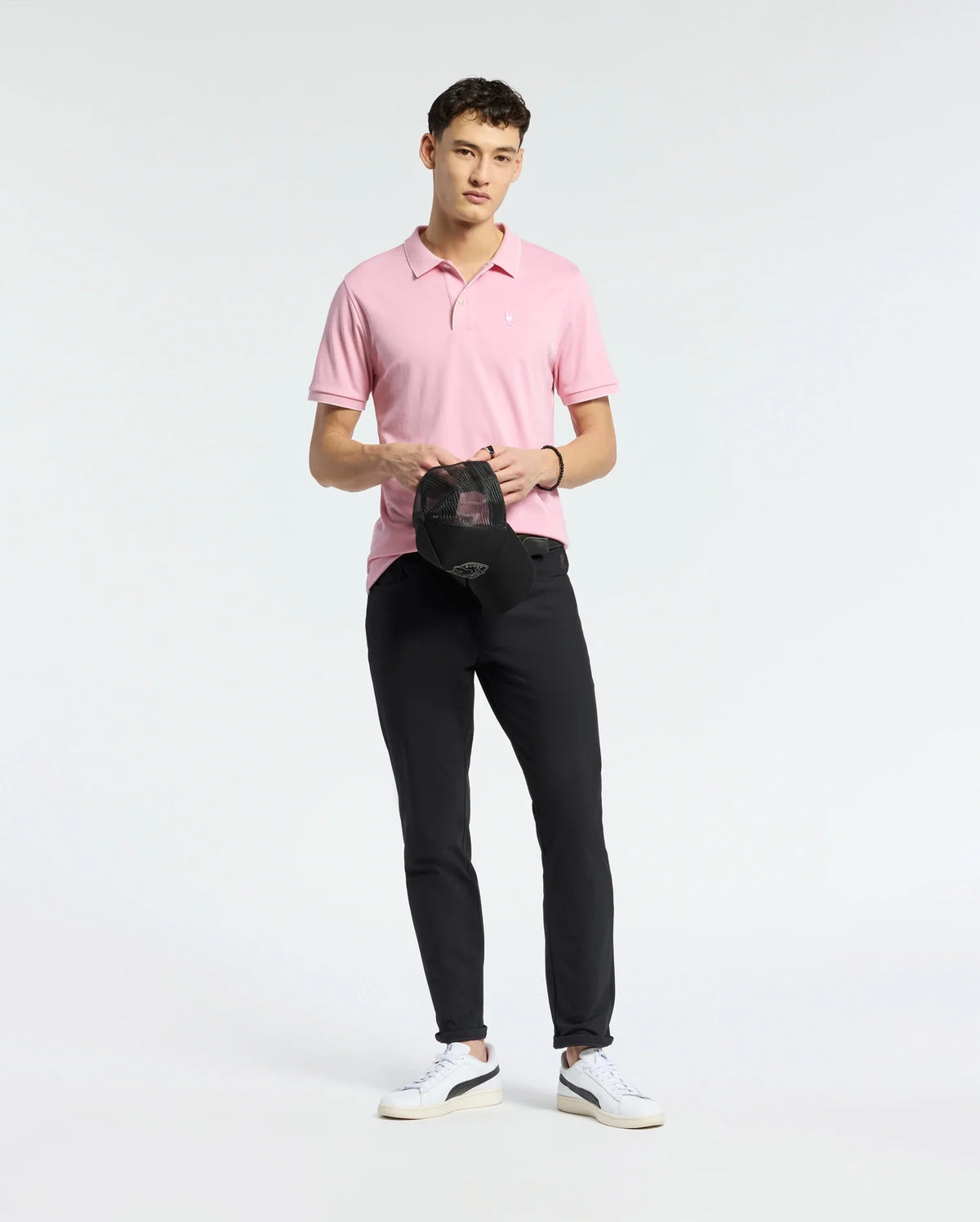 Wearing the Psycho Bunny MENS BRYCE PERFORMANCE PIQUE POLO in pink, paired with black pants and white sneakers, a person stands against a plain background. Holding a black cap with a neutral expression, they exude comfort and style in moisture-wicking attire.