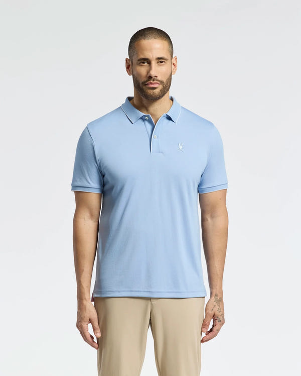 A bearded man stands against a white background wearing the Psycho Bunny MENS BRYCE PERFORMANCE PIQUE POLO in light blue, with a small white chest emblem, ensuring moisture wicking comfort. He pairs it with beige pants.