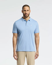 A bearded man stands against a white background wearing the Psycho Bunny MENS BRYCE PERFORMANCE PIQUE POLO in light blue, with a small white chest emblem, ensuring moisture wicking comfort. He pairs it with beige pants.