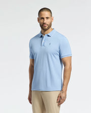 A man with a beard wears a light blue Psycho Bunny MENS BRYCE PERFORMANCE PIQUE POLO, B6K486F200, crafted from moisture-wicking fabric and featuring a chest logo for quick-dry comfort. He pairs it with beige pants against a plain white backdrop.