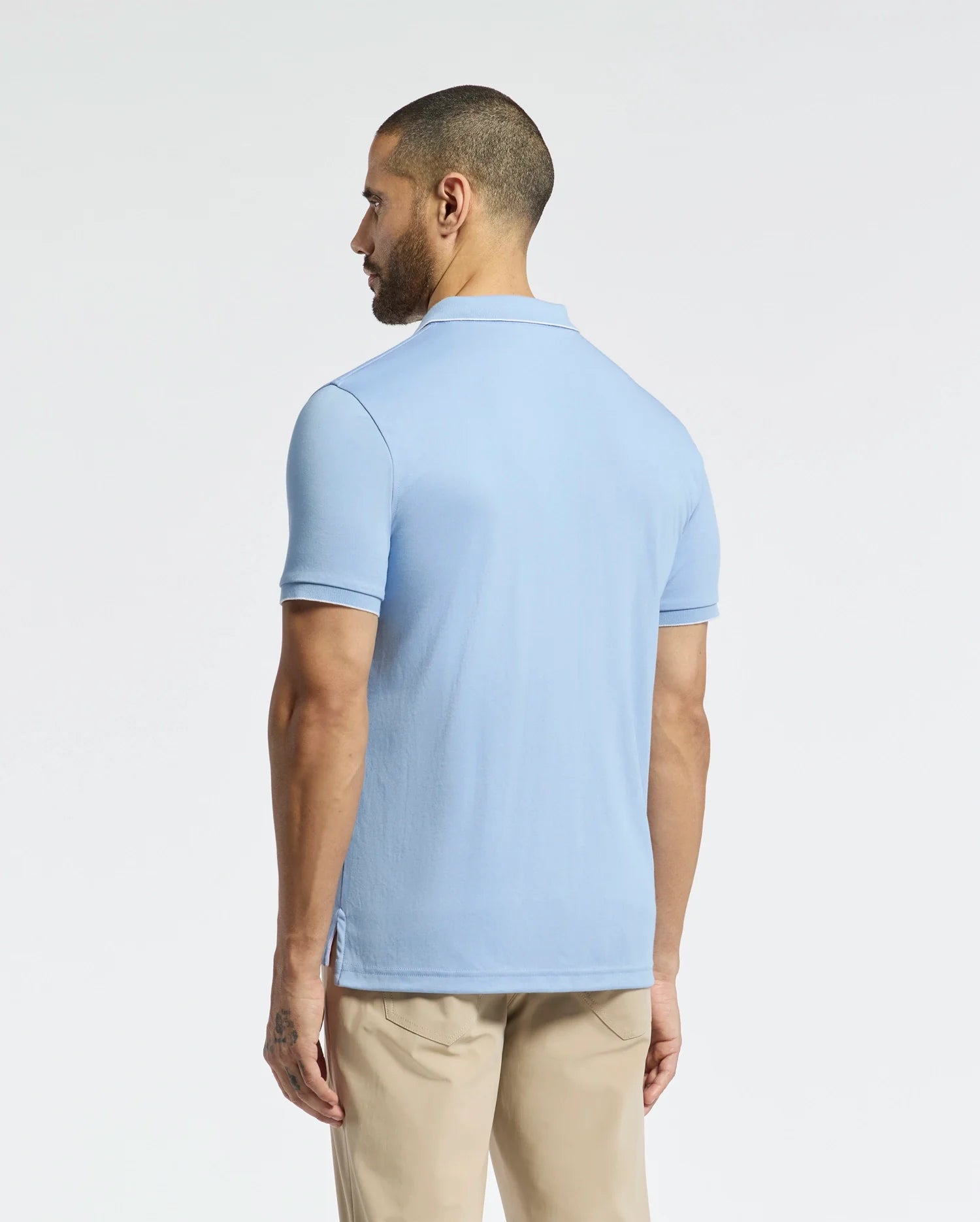 A man with short hair faces away, wearing a Psycho Bunny MENS BRYCE PERFORMANCE PIQUE POLO in light blue and beige pants, set against a plain white background.