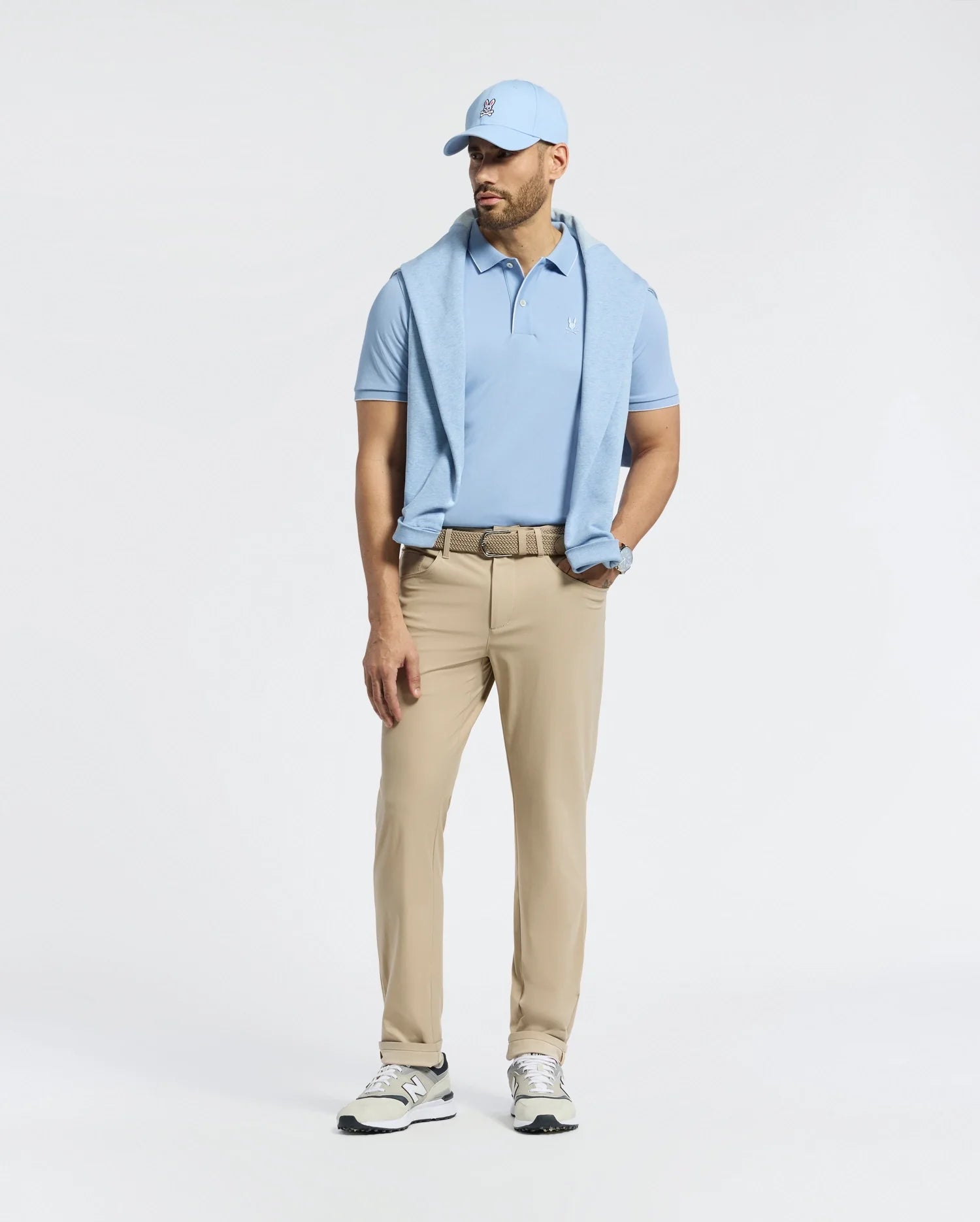 A man stands against a plain white background wearing a Psycho Bunny MENS BRYCE PERFORMANCE PIQUE POLO in light blue, with a matching cap and beige pants. He has a sweater over his shoulders, sports sneakers, wears a belt, and has a watch on his left wrist. The quick-dry fabric ensures comfort.