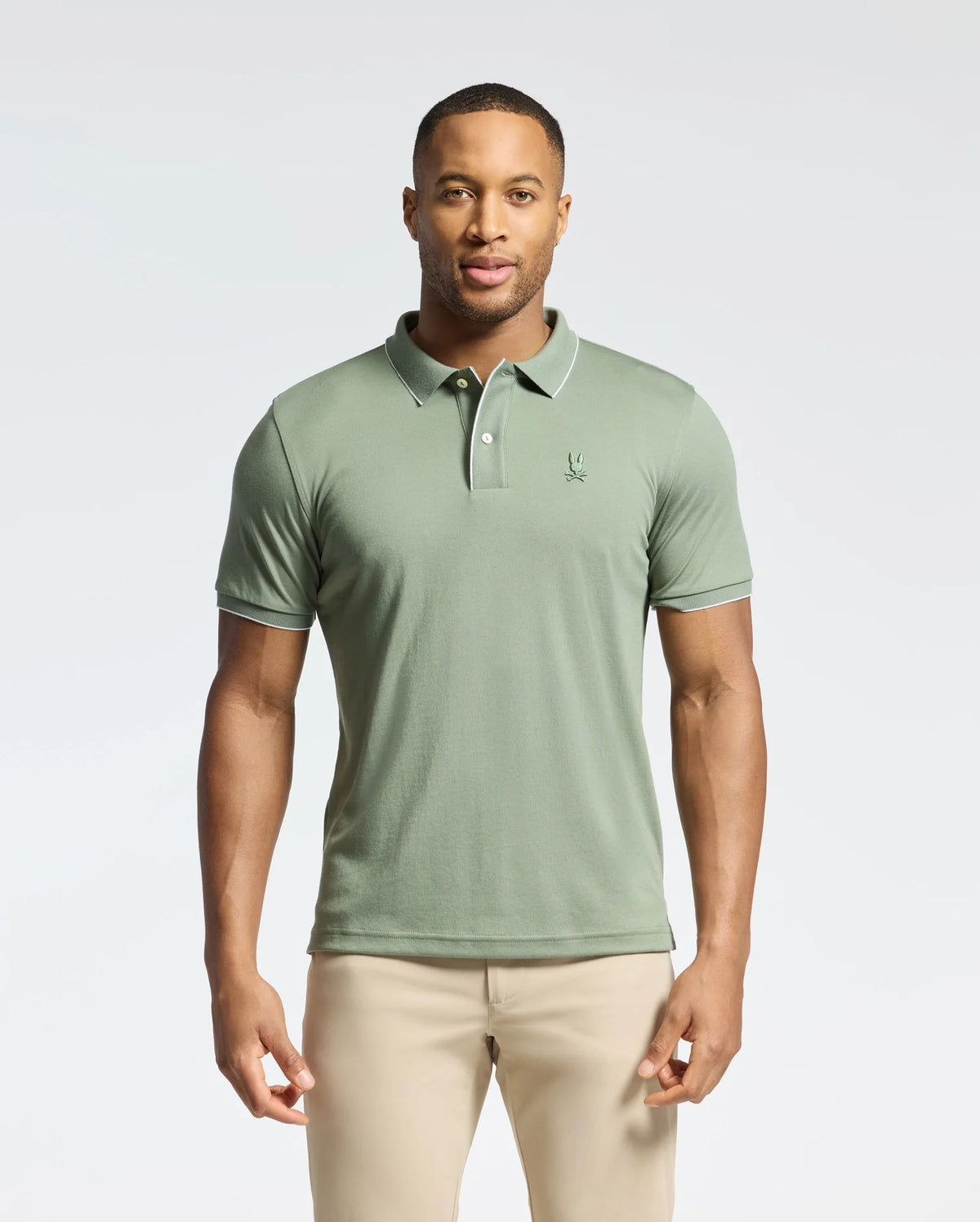 A man stands against a plain background wearing the Psycho Bunny MENS BRYCE PERFORMANCE PIQUE POLO (B6K486F200) in light green, made from breathable moisture-wicking fabric with a small embroidered logo on the chest, paired with beige pants.
