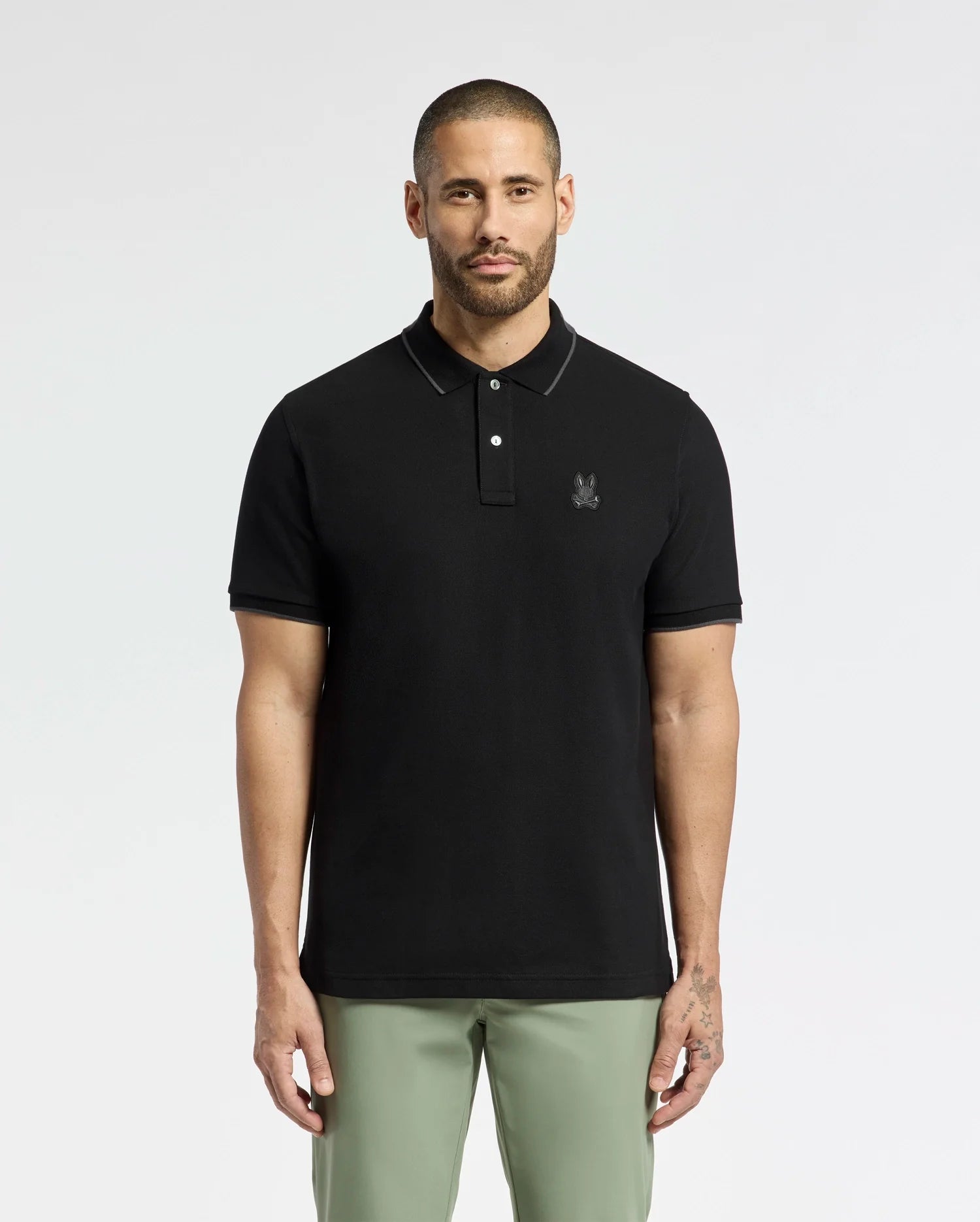 A man with short hair and a beard stands against a white background wearing the Psycho Bunny MENS NEW CASTLE PIQUE POLO (B6K487F200), a slim fit black polo with an embroidered bunny patch on the chest. He pairs it with light green pants for a laid-back style.