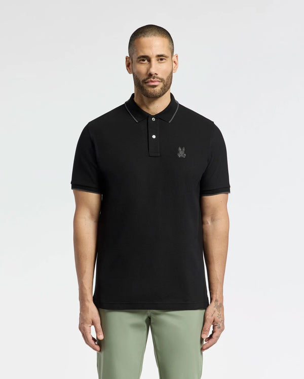 A man with short hair and a beard stands against a white background wearing the Psycho Bunny MENS NEW CASTLE PIQUE POLO (B6K487F200), a slim fit black polo with an embroidered bunny patch on the chest. He pairs it with light green pants for a laid-back style.