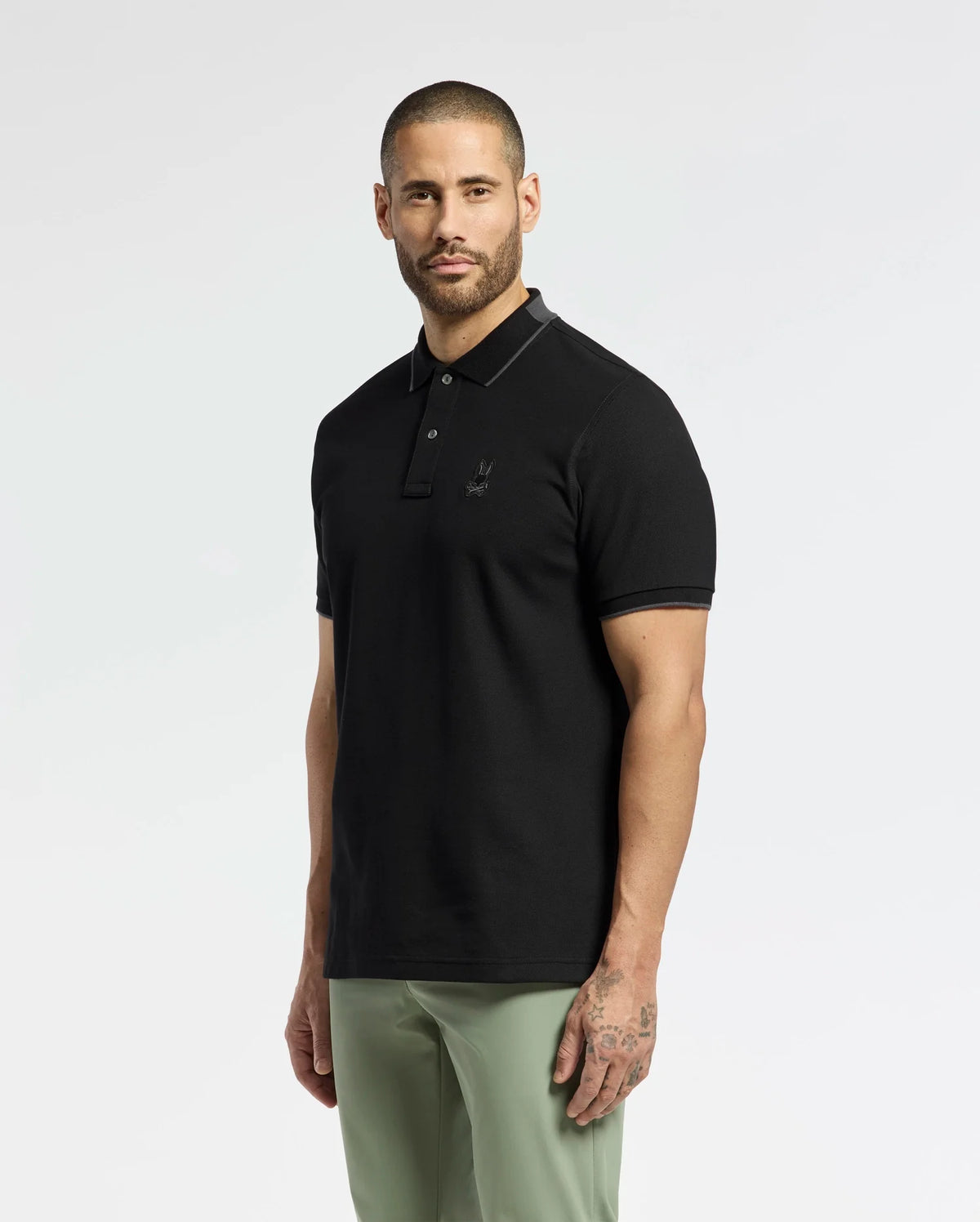 A bearded man with short hair wears a Psycho Bunny MENS NEW CASTLE PIQUE POLO (B6K487F200) in black, made of soft Pima cotton with an embroidered bunny patch. His slim-fit, light green pants reveal tattoos on his left arm against a plain white background.