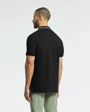 A man with short hair and a beard turns away, showcasing the sleek back of the Psycho Bunny MENS NEW CASTLE PIQUE POLO - B6K487F200 in black with a dark grey collar. Paired with light green pants, this pima cotton polo offers comfort and elegance against a plain white backdrop.