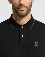A man dons the Psycho Bunny MENS NEW CASTLE PIQUE POLO (B6K487F200), a slim fit black polo made from Pima cotton. It features a buttoned placket and an embroidered chest bunny patch on the left, all set against a plain gray background, showcasing its design with the focus away from his partially visible face.