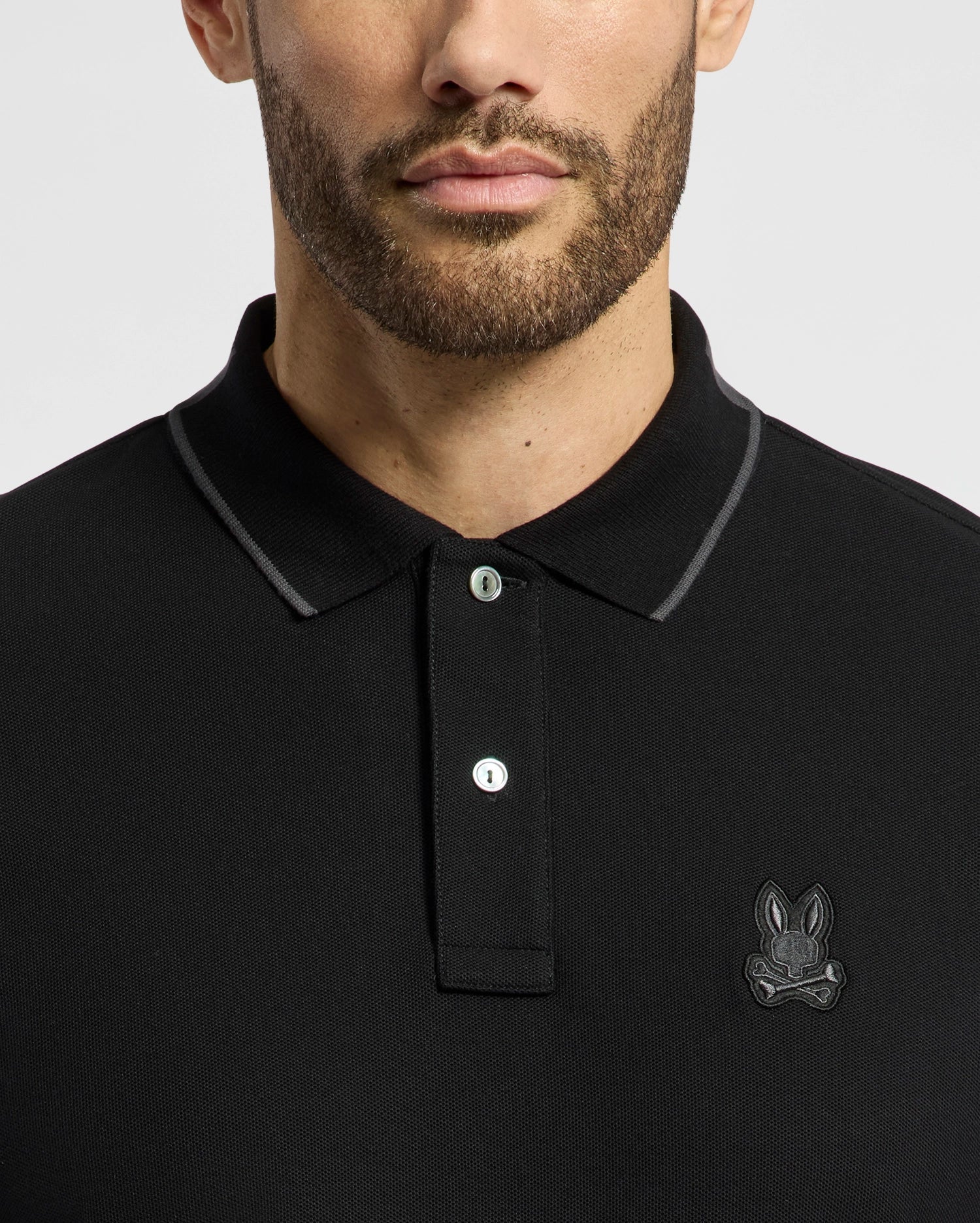 A man dons the Psycho Bunny MENS NEW CASTLE PIQUE POLO (B6K487F200), a slim fit black polo made from Pima cotton. It features a buttoned placket and an embroidered chest bunny patch on the left, all set against a plain gray background, showcasing its design with the focus away from his partially visible face.