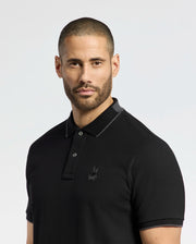 A bearded man with short hair wears the Psycho Bunny MENS NEW CASTLE PIQUE POLO (B6K487F200), a slim-fit black polo in luxurious Pima cotton with a subtle chest bunny patch against a plain light background.