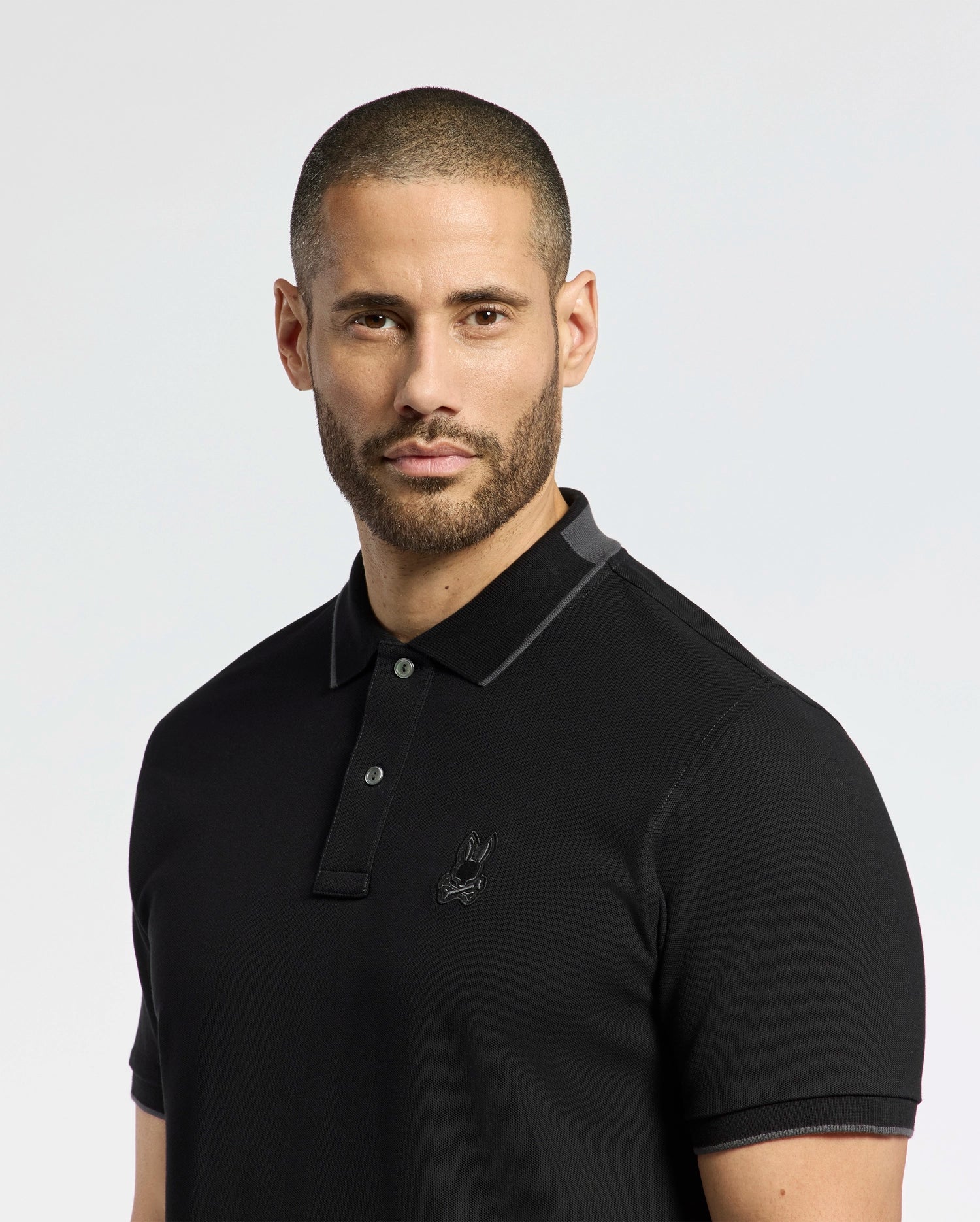 A bearded man with short hair wears the Psycho Bunny MENS NEW CASTLE PIQUE POLO (B6K487F200), a slim-fit black polo in luxurious Pima cotton with a subtle chest bunny patch against a plain light background.
