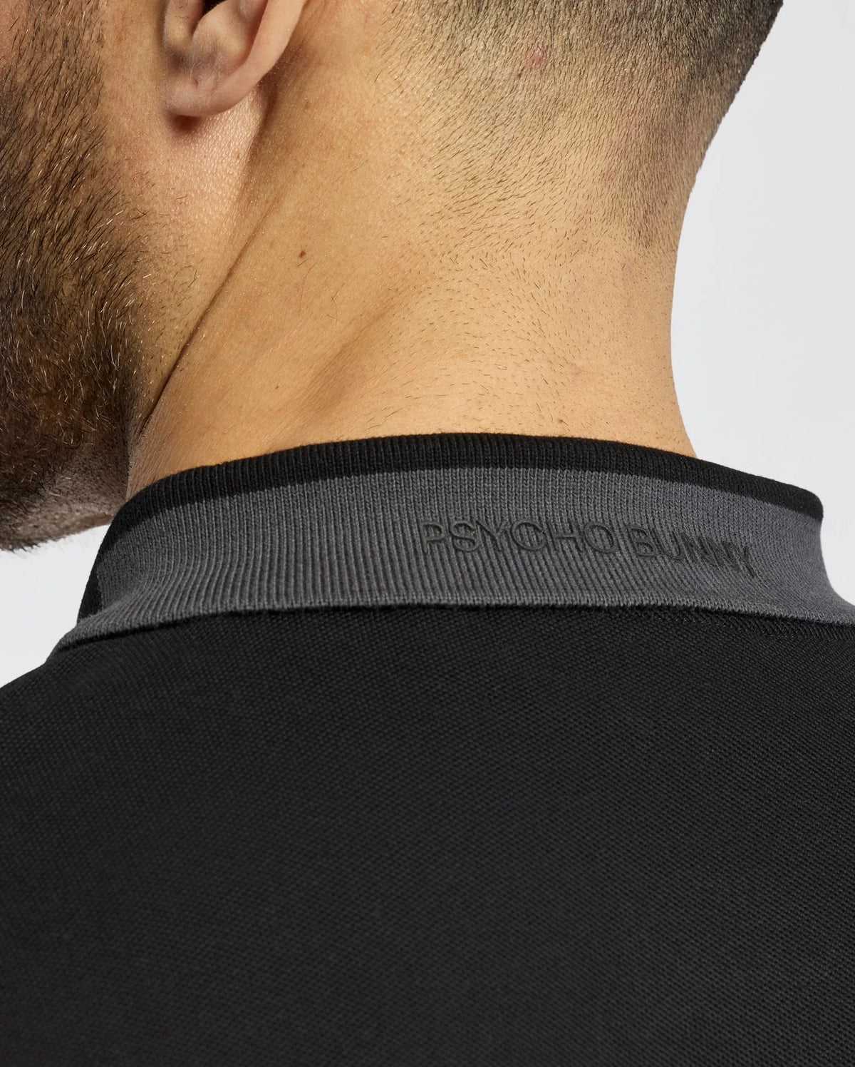 Close-up of the back of a person’s neck in a Psycho Bunny slim-fit black MENS NEW CASTLE PIQUE POLO, B6K487F200. "PSYCHO BUNNY" is subtly embossed on the gray collar. Crafted from soft Pima cotton, it features a sleek plain gray background.