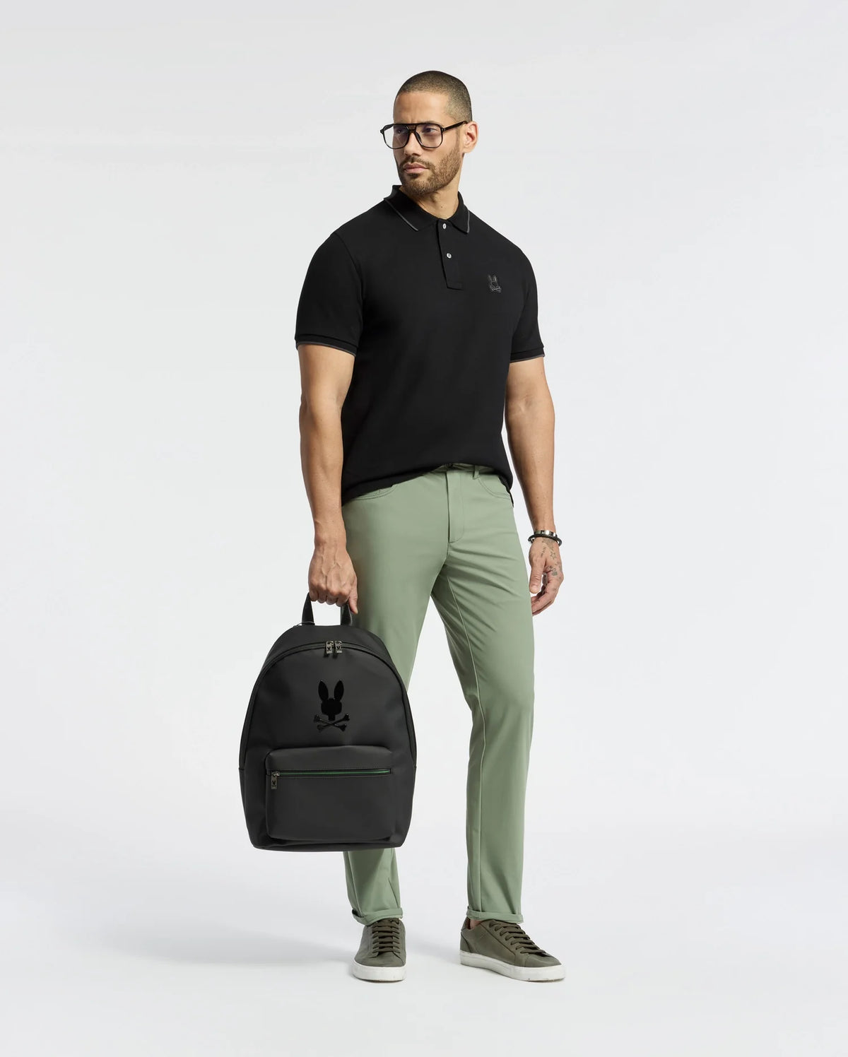 A man in a Psycho Bunny MENS NEW CASTLE PIQUE POLO - B6K487F200, black slim-fit with an embroidered bunny chest patch, stands holding a black backpack. He pairs it with green pants, gray sneakers, and glasses against a plain white background.
