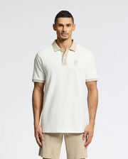 A man wears a Psycho Bunny beige MENS NEW CASTLE PIQUE POLO - B6K487F200 and shorts against a plain background. The 100% Pima Cotton polo features an embroidered chest Bunny patch logo, enhancing the charm of the casual neutral-toned outfit.