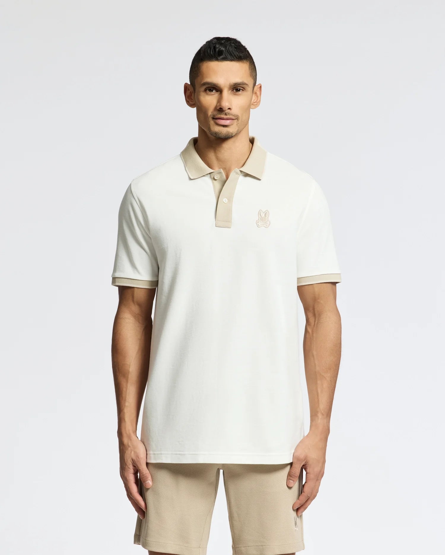 A man wears a Psycho Bunny beige MENS NEW CASTLE PIQUE POLO - B6K487F200 and shorts against a plain background. The 100% Pima Cotton polo features an embroidered chest Bunny patch logo, enhancing the charm of the casual neutral-toned outfit.