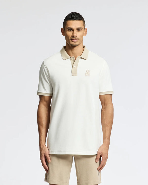 A man wears a Psycho Bunny beige MENS NEW CASTLE PIQUE POLO - B6K487F200 and shorts against a plain background. The 100% Pima Cotton polo features an embroidered chest Bunny patch logo, enhancing the charm of the casual neutral-toned outfit.