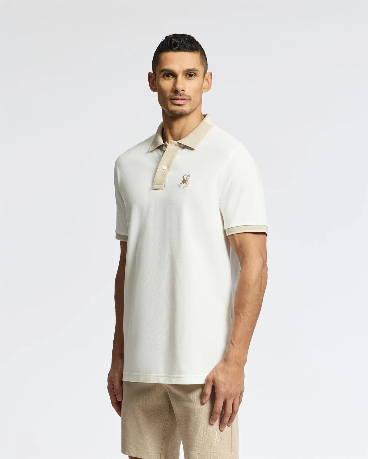A man sports a Psycho Bunny's Mens New Castle Pique Polo (B6K487F200), made from 100% Pima Cotton with a light brown collar and embroidered Bunny patch on the chest, set against a plain background. He pairs it with beige shorts, arms relaxed by his sides as he faces the camera.